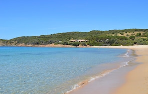 Ein Strand mit klarem Wasser, einem Sandufer und einem Gebäude mit Dach auf einem Hügel.
