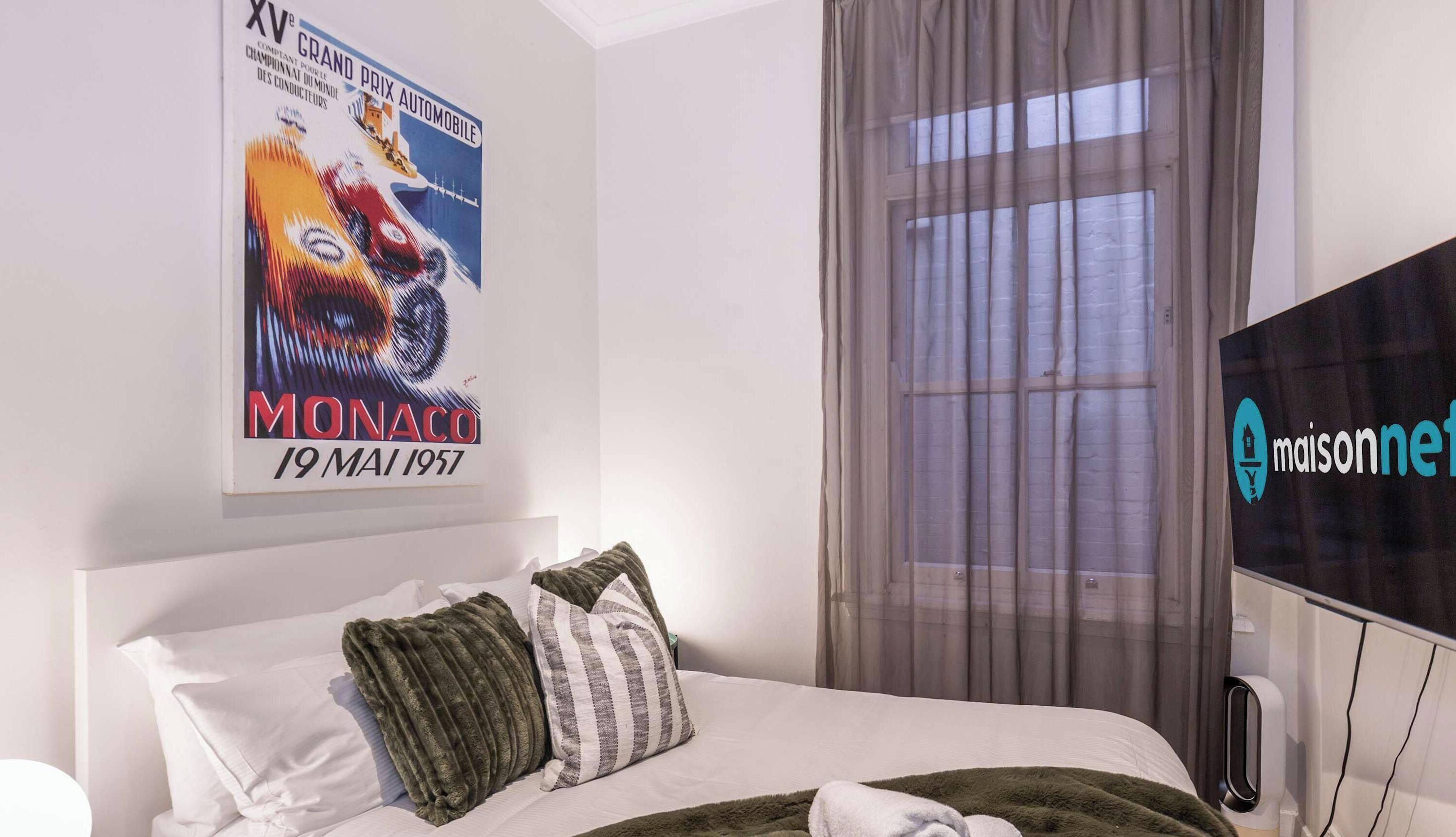 A hotel room with a bed, a poster on the wall, a window with curtains, and a television.
