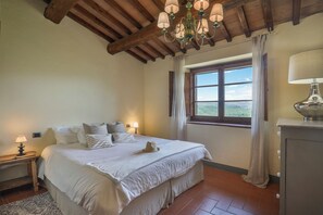 A bedroom with a large bed, a nightstand with a lamp, a window with curtains, and a chandelier. - Casa Poggio in Badia in the Chianti Shire (Toscana)