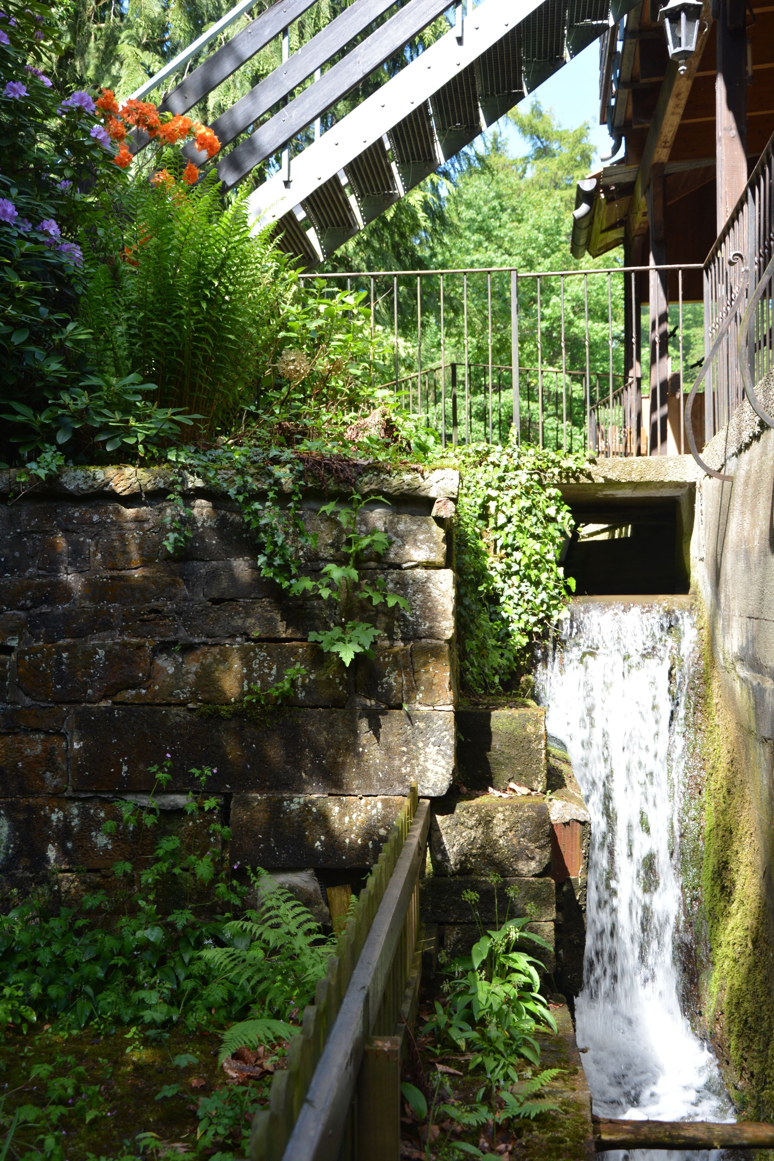 Eine steinerne Mauer mit einem kleinen Wasserfall, umgeben von üppigem Grün und einer Holzbrücke.