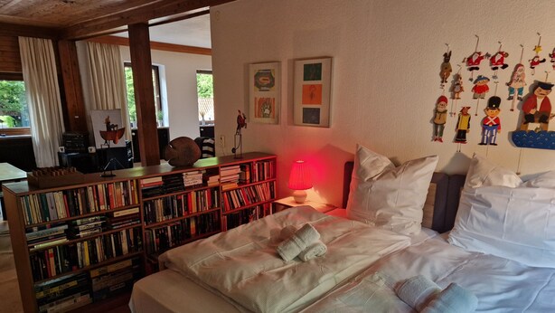 A bedroom with a large bed, a bookshelf, and a wall with hanging decorations.