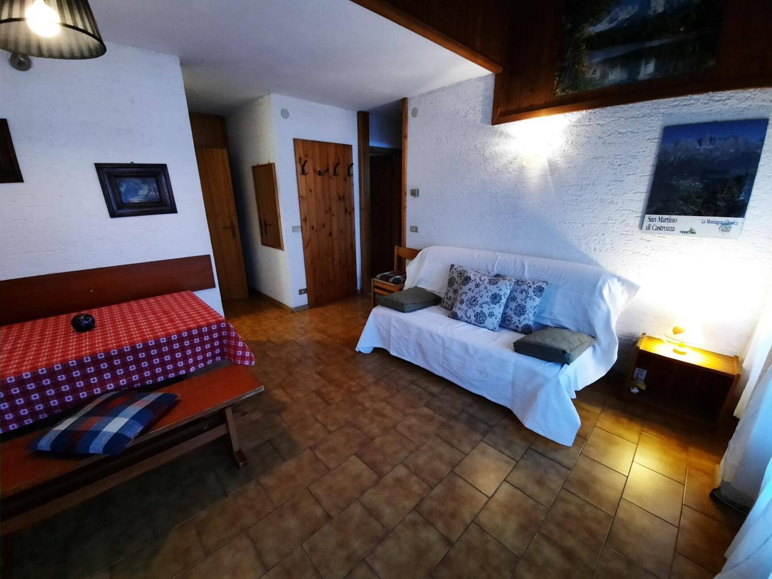 Una stanza con un letto, un divano, un tavolo con tovaglia a quadri rossi e una lampada fissata al muro.