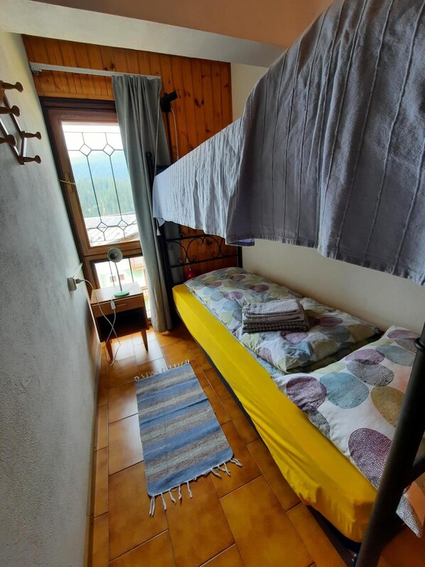A small, cozy room with a bunk bed, a window with curtains, a wooden floor, and a striped rug. - Apartment 'Stregatto Dalla Luna - Dolomiti' with Mountain View, Private Terrace and Wi-Fi (San Martino di Castrozza)