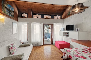 Une pièce chaleureuse avec un plafond en bois, un canapé, une petite table, un téléviseur et une vue sur un balcon équipé d’une chaise.
