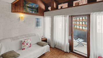 Ett rum med ett träpanelat tak, en vit, strukturerad vägg, en soffa med kuddar, ett litet bord med en lampa, ett fönster med gardiner och utsikt över en byggnad utanför.