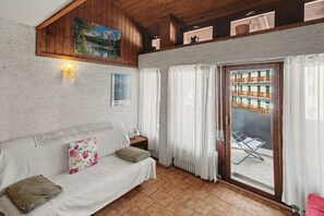 A room with a wooden ceiling, a white textured wall, a sofa with cushions, a small table with a lamp, a window with curtains, and a view of a building outside.