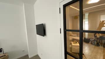 A modern interior with a flat-screen TV mounted on a white wall, a wooden side table, and a sliding glass door leading to a living area with a sofa and a coffee table.