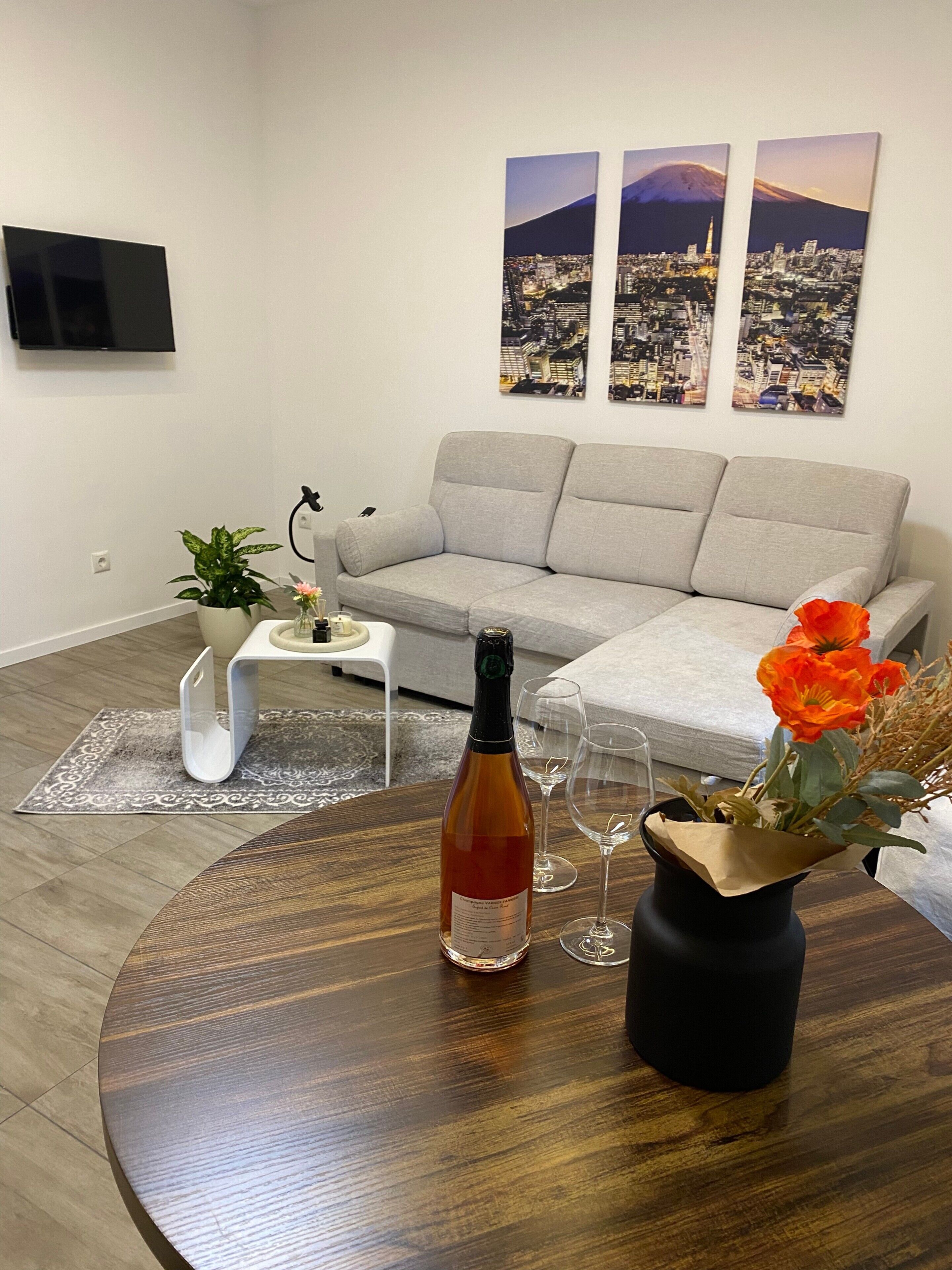 A modern living room with a grey sofa, a round wooden coffee table, a bottle of wine, and a vase with flowers. There are three framed cityscape pictures on the wall.