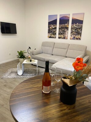 A modern living room with a grey sofa, a round wooden coffee table, a bottle of wine, and a vase with flowers. There are three framed cityscape pictures on the wall. - Apartment near Bonn & Ahr Valley – Modern Vacation Rental in Grafschaft (Grafschaft)