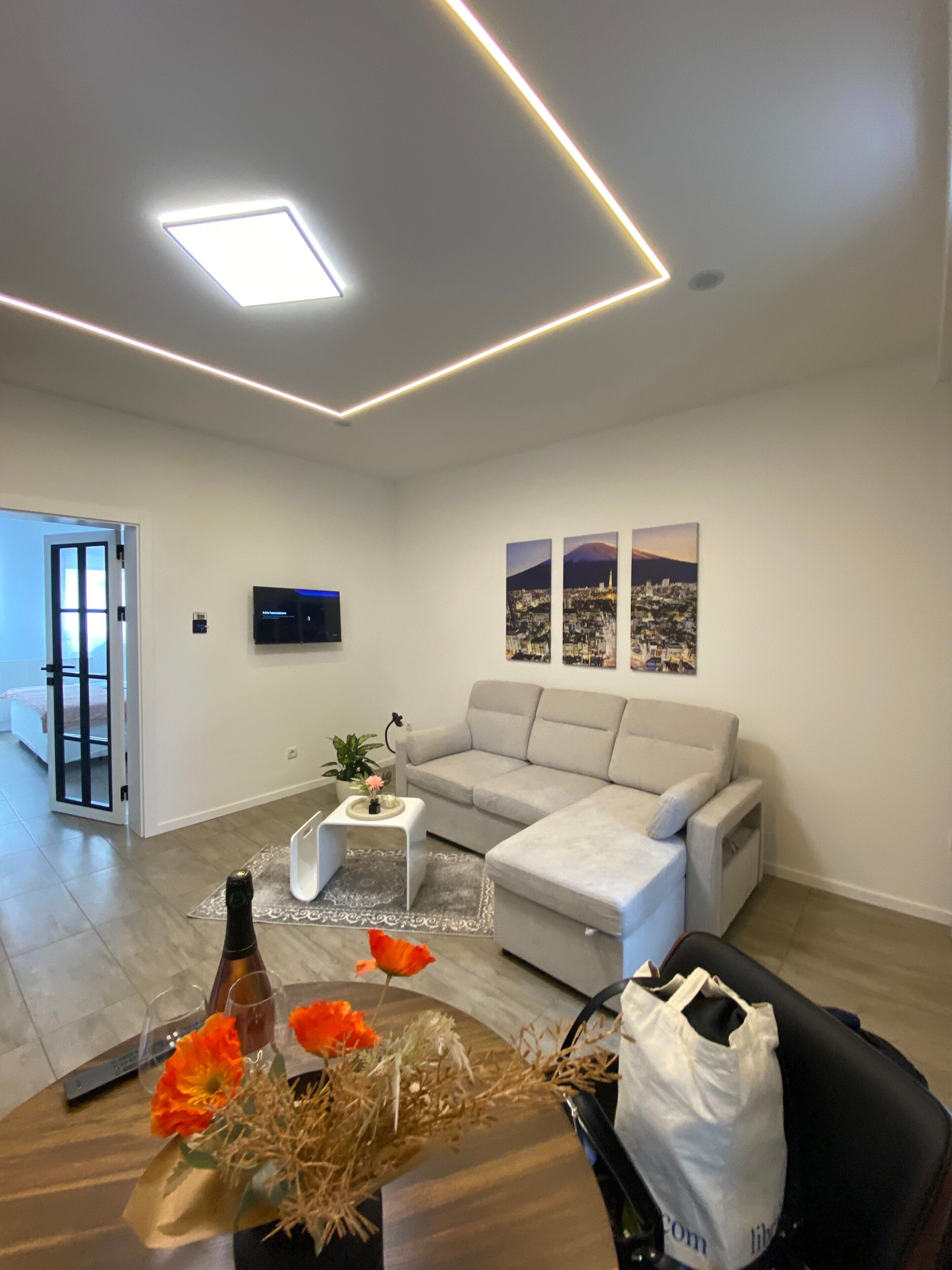 A modern living room with a sofa, a coffee table, and a flat-screen TV mounted on the wall. There are three framed pictures on the wall, and a door leading to another room.