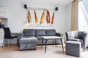 A modern living room with a grey sofa, a small table, and a chair. There is a painting on the wall and a kitchen area in the background. - Dünenvilla 10, Apartment 04 (Lübeck-Travemünde)