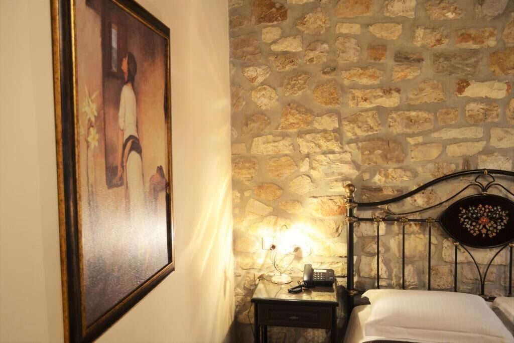 A room with a stone wall, a bed with a headboard, a bedside table with a lamp, and a framed picture on the wall.