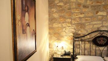 A room with a stone wall, a bed with a headboard, a bedside table with a lamp, and a framed picture on the wall.