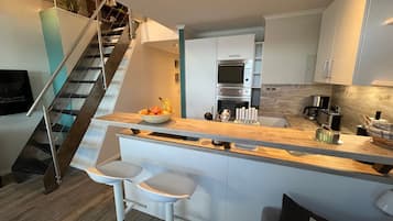 Una cocina moderna con una isla central, taburetes de bar, horno empotrado y una escalera que lleva a un nivel superior.