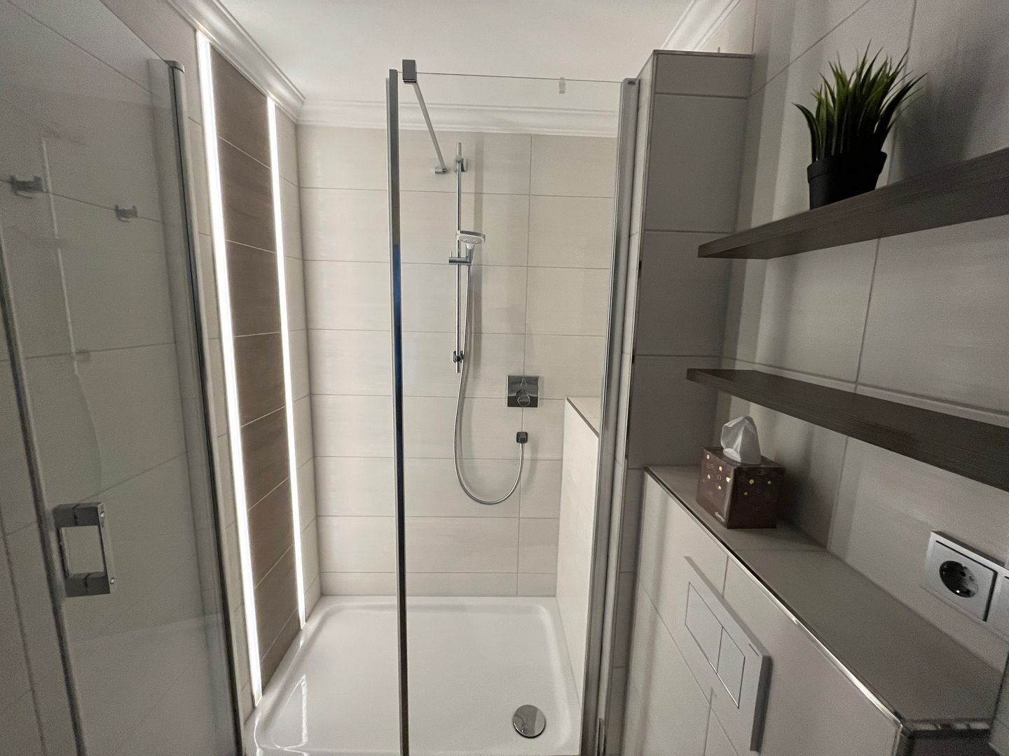 Ein modernes Badezimmer mit einer in Glas eingefassten Dusche, weißen Fliesenwänden und einer eingebauten Regaleinrichtung.