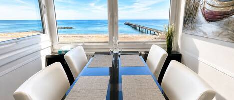 A dining area with a view of the beach and pier, featuring a table with four chairs and a painting on the wall.