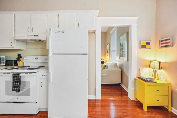 A kitchen with white cabinets, a stove, and a refrigerator. There is a yellow bedside table with a lamp and a bed in the adjacent room. - Historical Townhouse on UT campus | Free Parking | Minutes to Downtown ❤️ (Austin)