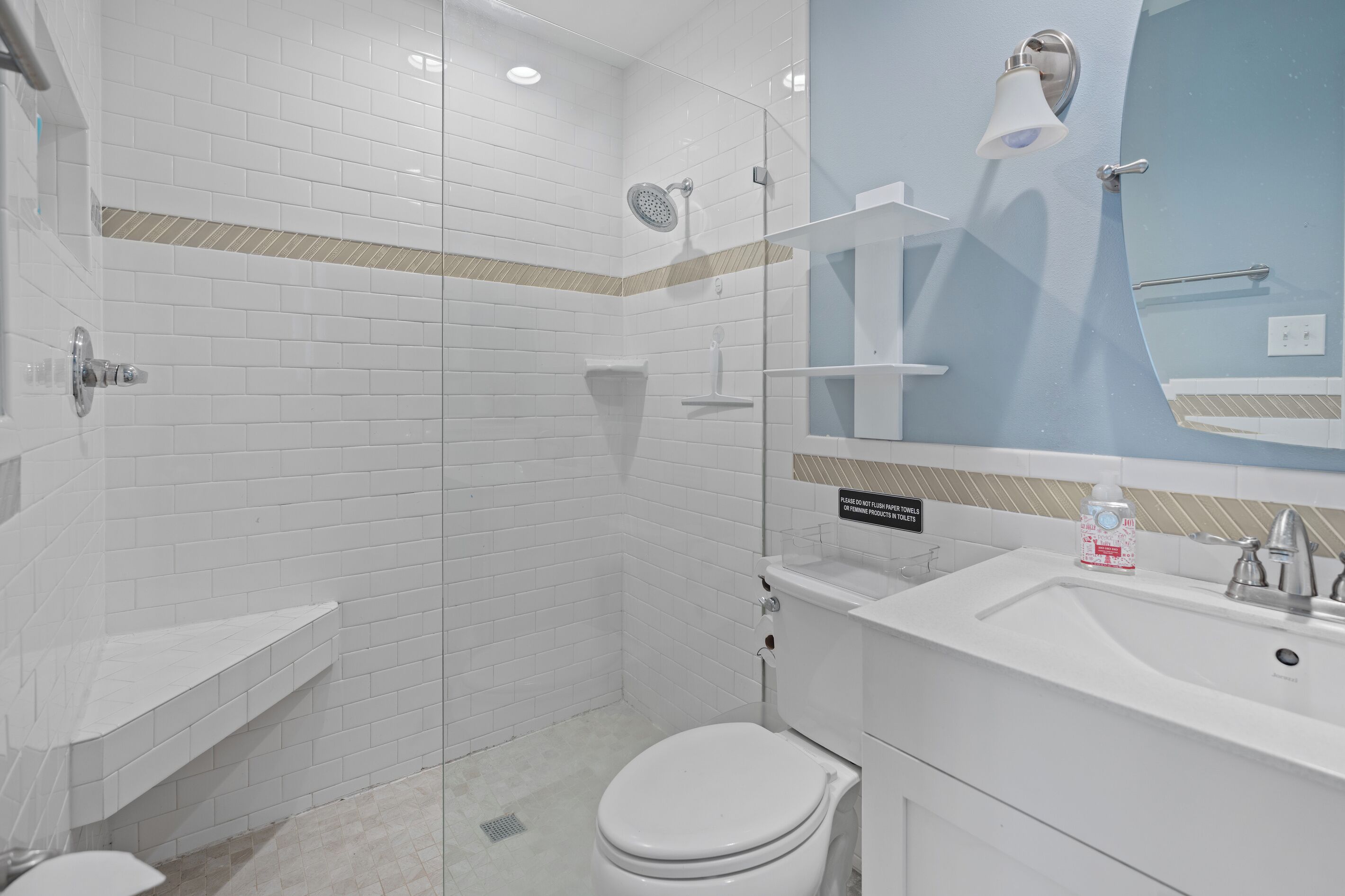 Ein modernes Badezimmer mit weißen Fliesen im U-Bahn-Stil, einer Glasdusche, einer Toilette, einem Waschbecken mit Spiegel und einem Schild, das darauf hinweist, keine Papierhandtücher oder Hygieneartikel auszuspülen.