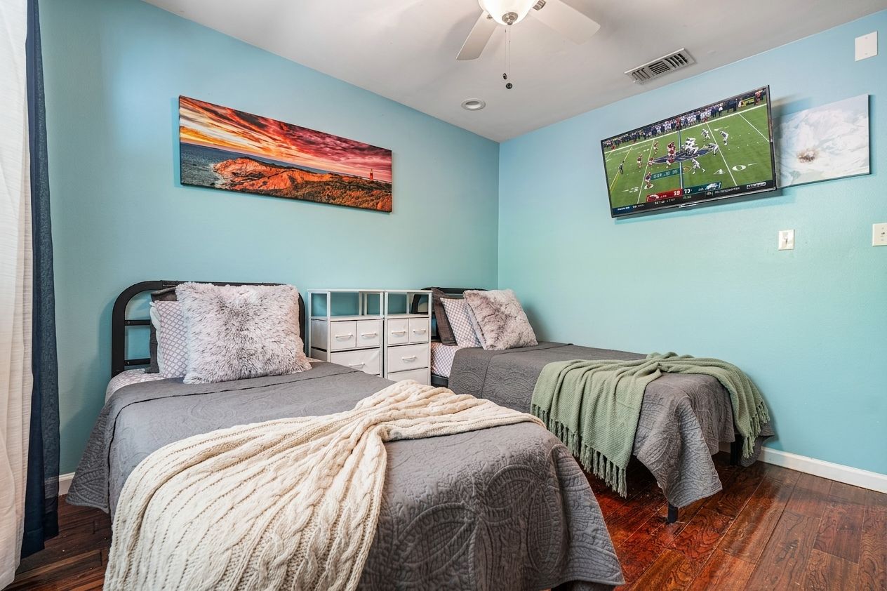 Ein Schlafzimmer mit zwei Betten, einem Deckenventilator, einem an der Wand befestigten Fernseher und Kunstwerken.