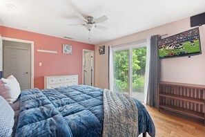 Quarto com cama, ventoinha no teto, TV fixada na parede e porta de vidro deslizante para uma varanda.