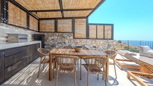 An outdoor patio with a stone wall, a wooden table, wicker chairs, and a view of the sea. - Metatulia Villa 7 – Private Pool, Jacuzzi & Views (Plakias)