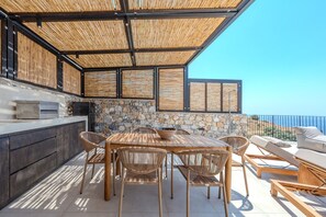 Eine Terrasse im Freien mit einer Steinsäule, einem Holztisch, Korbstühlen und Blick aufs Meer.