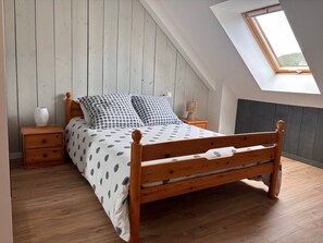 A bedroom with a sloped ceiling, a wooden bed frame, a nightstand, a lamp, and a window with a view of greenery. - Holiday Home 'Gîte 6 Personnes' with Private Terrace, Private Garden and Wi-Fi (Heugueville-sur-Sienne)