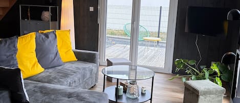 Een moderne woonkamer met een grijze bank, gele kussens, een glazen salontafel, een houten vloer, een trap en een schuifdeur van glas die leidt naar een buitenruimte.