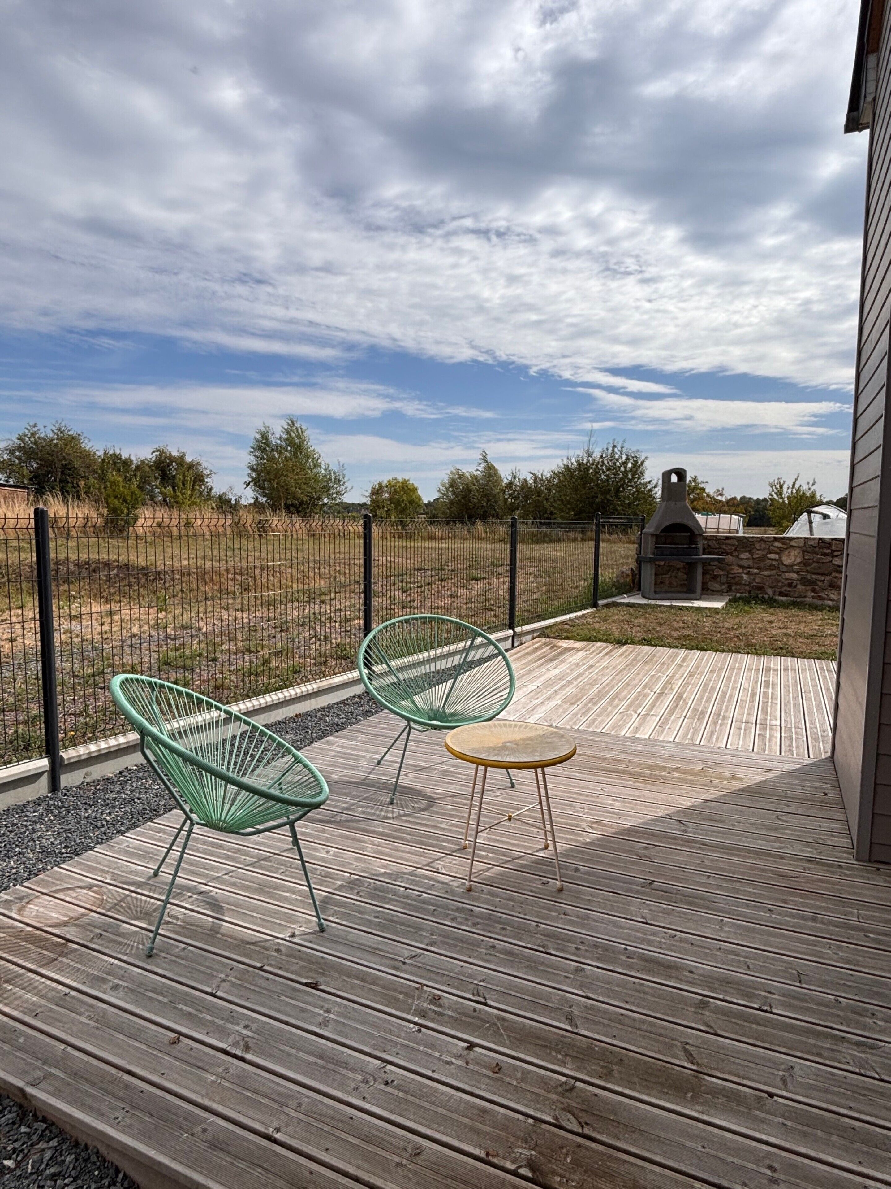 Un patio exterior con dos sillas verdes y una mesita, una parrilla de piedra y vistas a un área cercada con árboles.