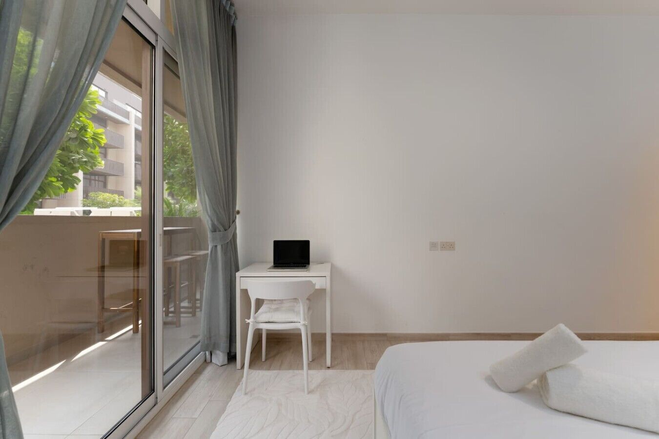 A bedroom with a bed, a desk with a laptop, a chair, and a large window with curtains.