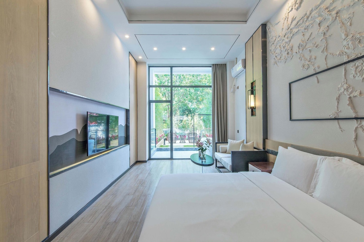 A modern hotel room with a large bed, a TV mounted on the wall, a small table, and a view of the outdoors through a glass door.