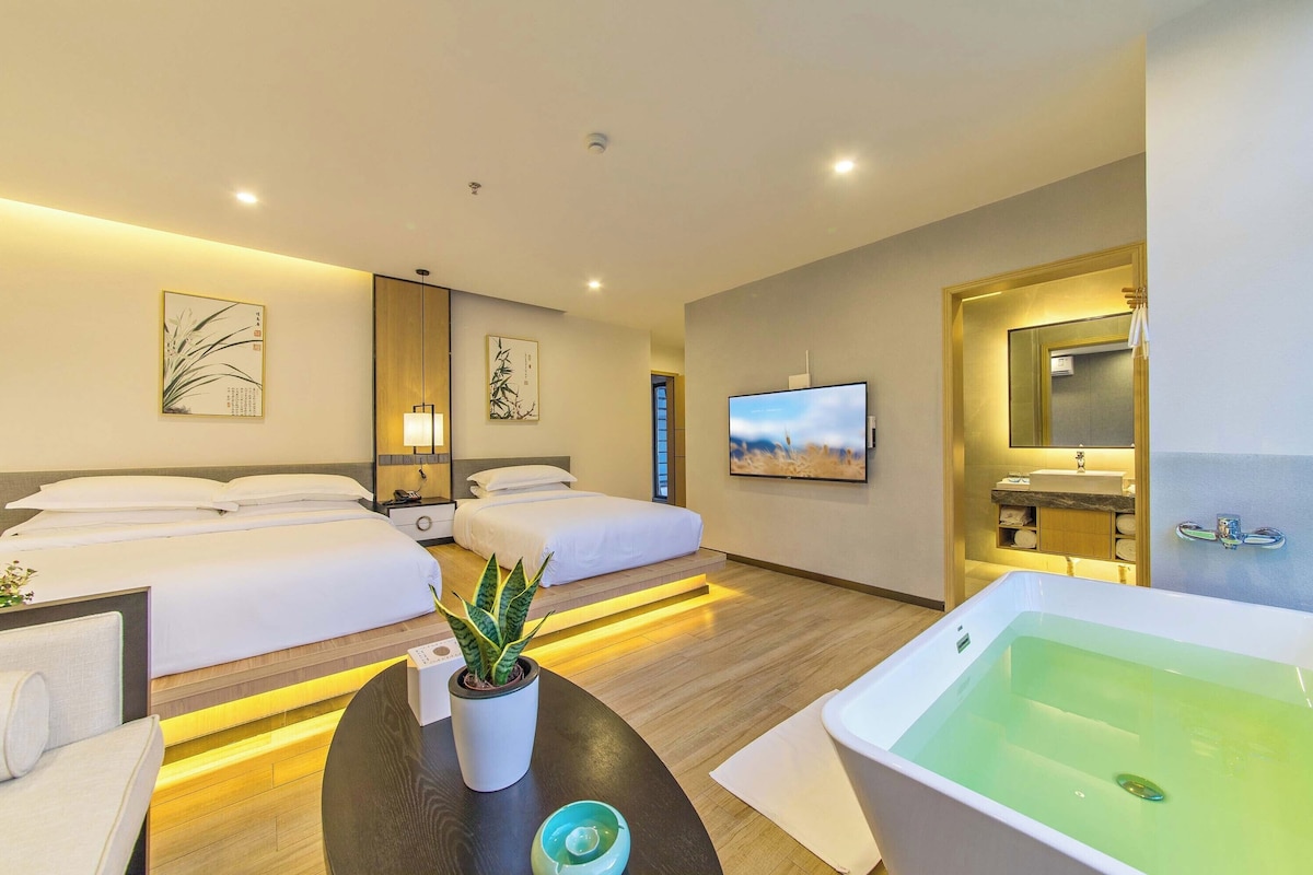 A hotel room with two beds, a large window, a TV, a bathtub, and artwork on the walls.