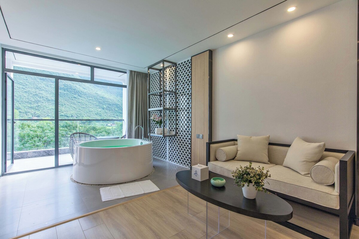 A modern living room with a sofa, a round bathtub, and a view of a mountainous landscape through large windows.