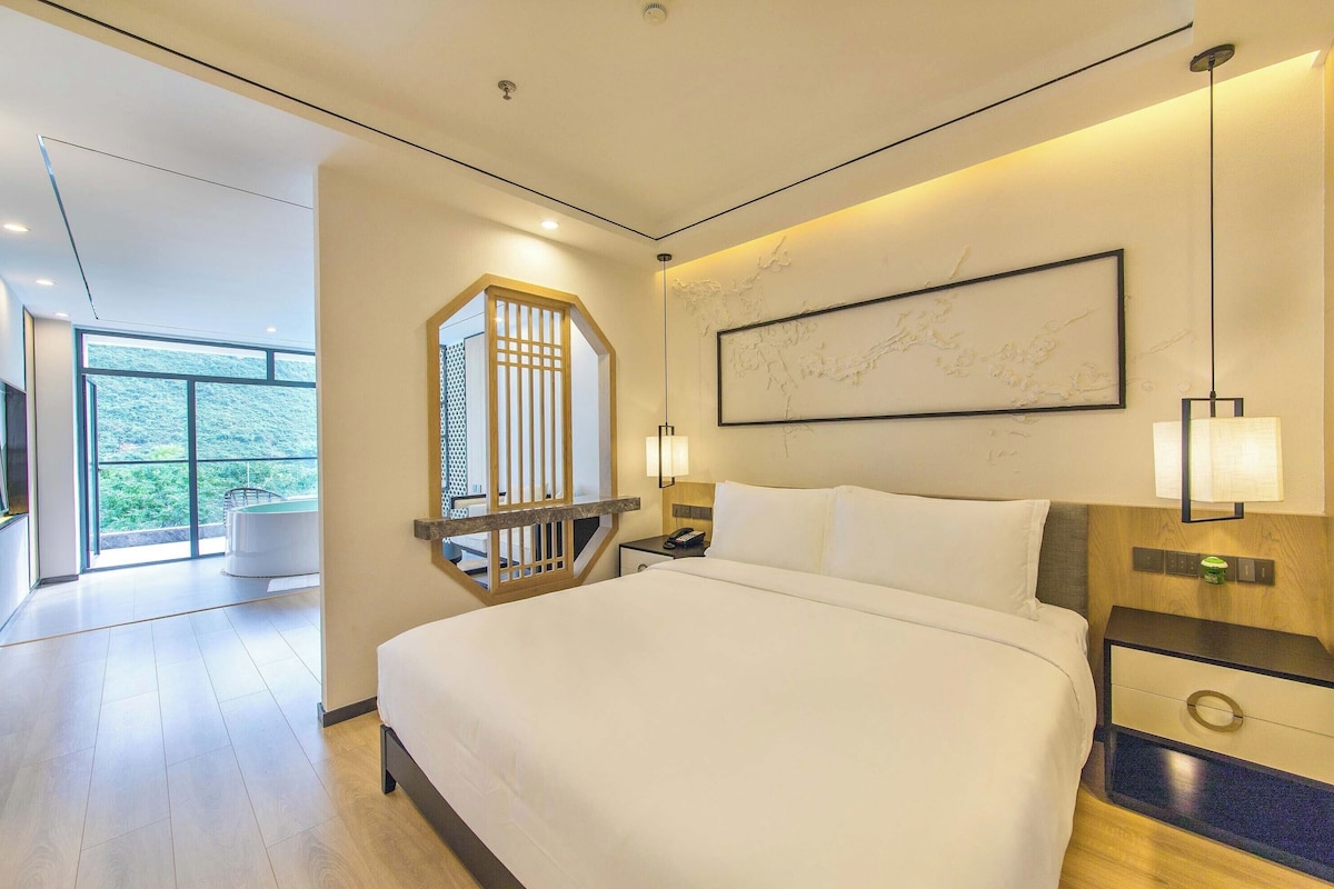 A modern hotel room with a large bed, a desk with a lamp, a view of the outdoors, and a large window.