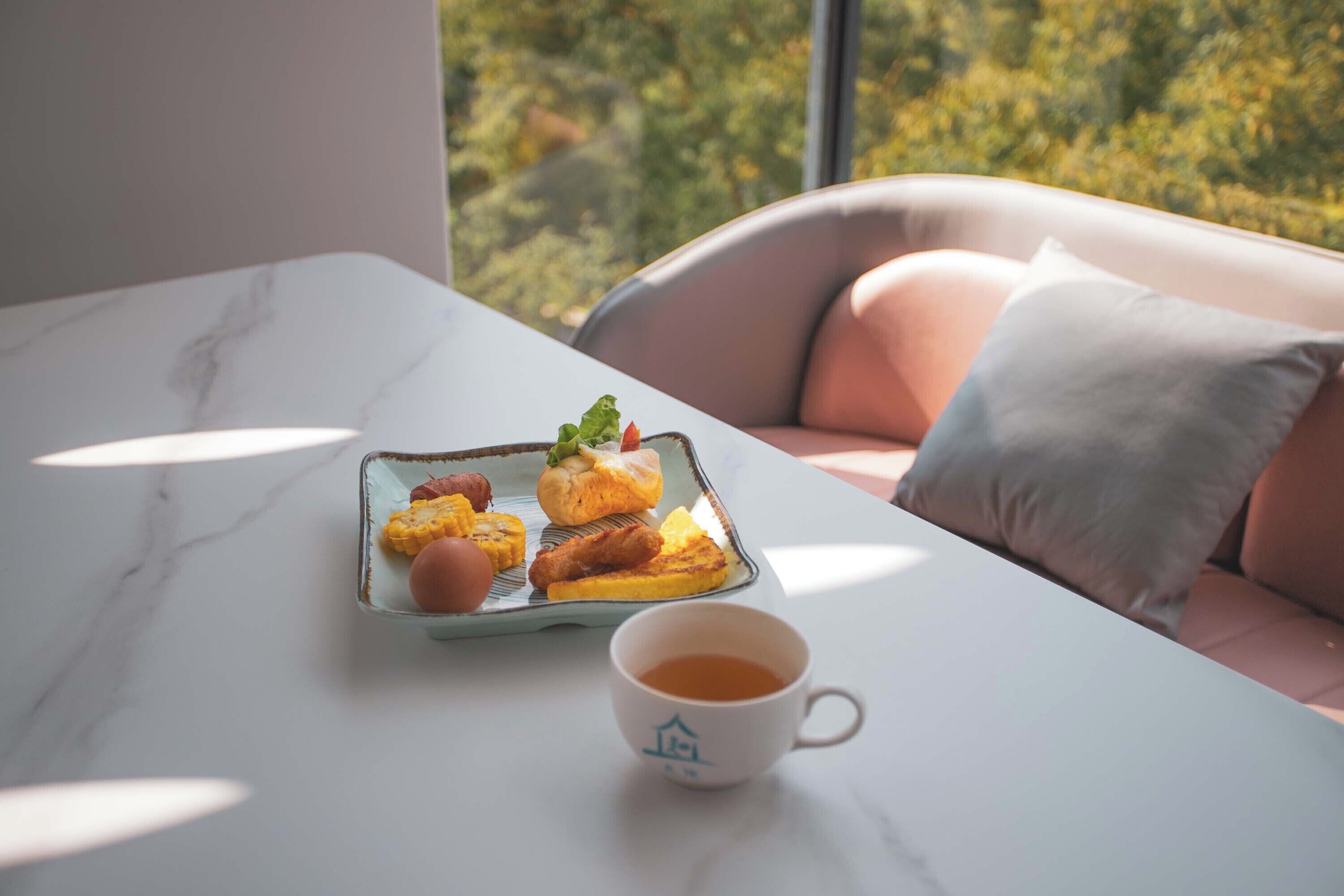 A table with a tray of food and a cup of coffee, a cushioned chair, and a view of greenery through a window.