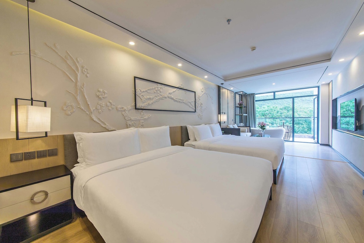 A modern hotel room with two beds, a large window, and a view of greenery.
