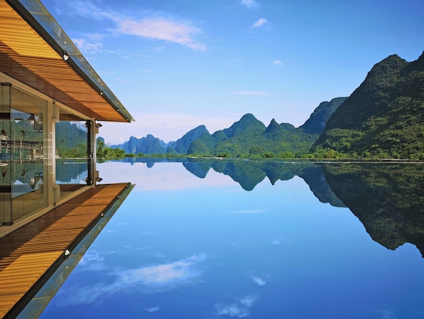 A modern building with a glass facade and wooden deck overlooking a calm lake with mountain reflections. - Liangsu Shili Jiangshan Hotel (Guilin)