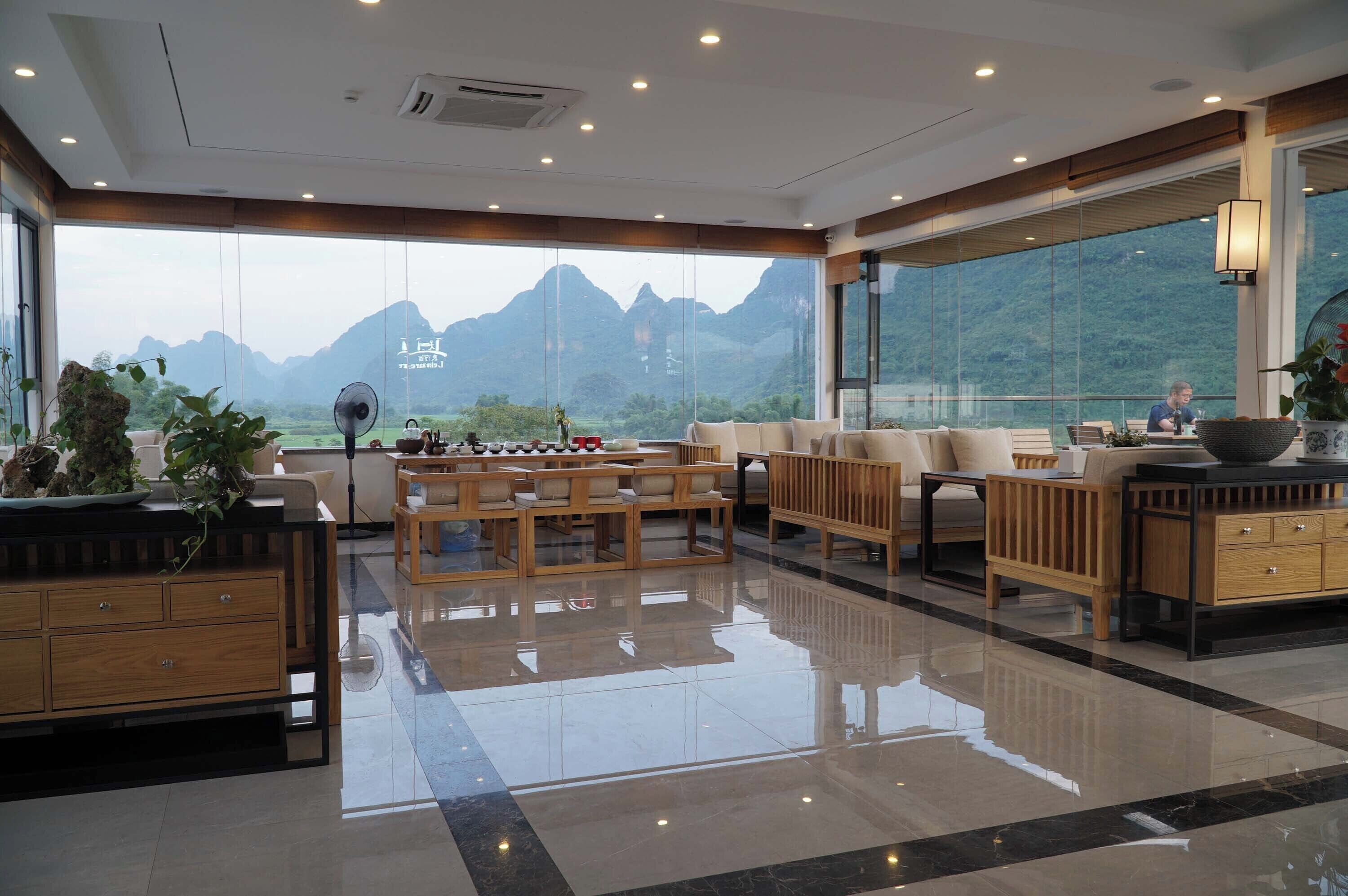 A spacious living area with a view of mountains through large windows, wooden furniture, and a polished floor.
