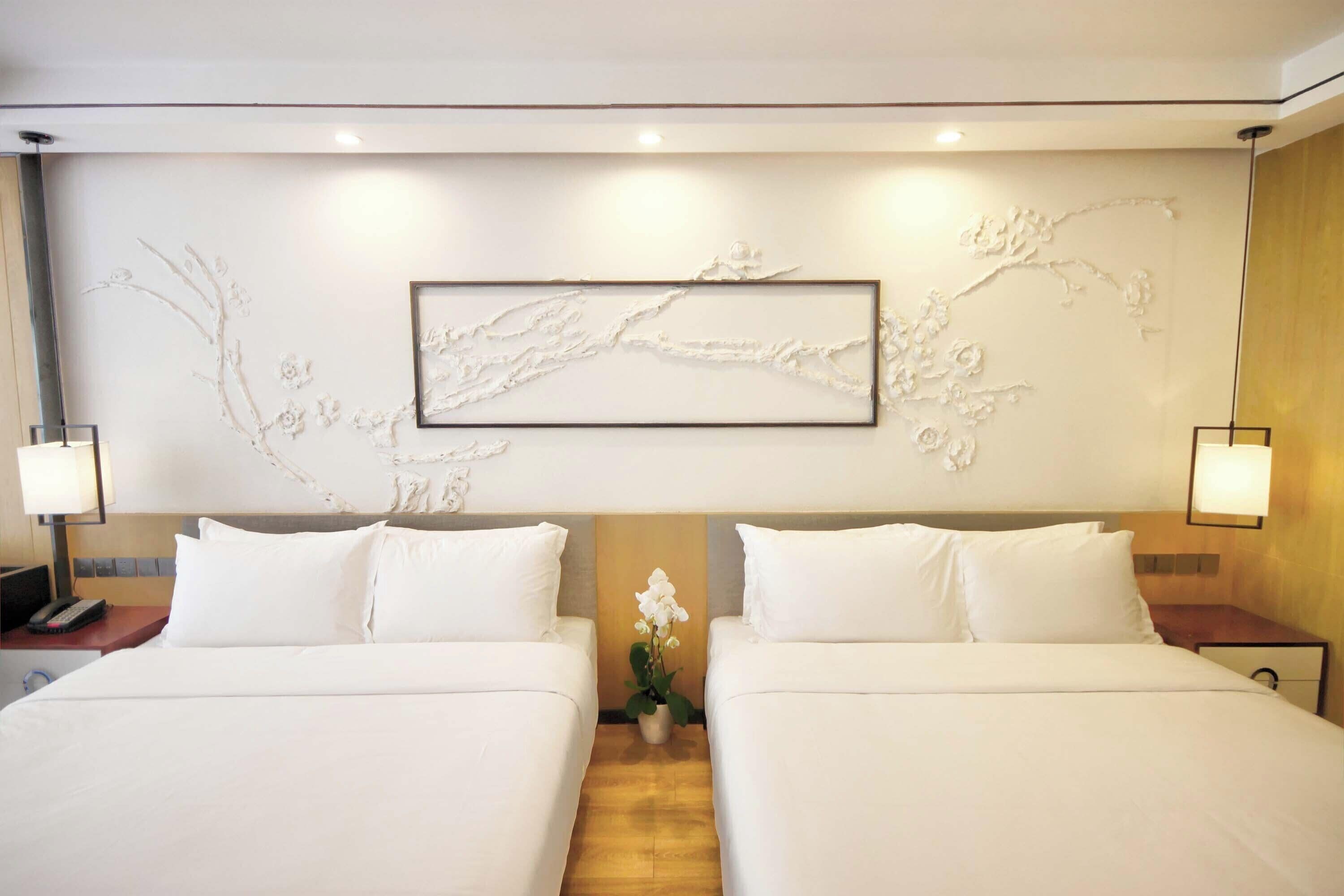 A hotel room with two beds, a wall-mounted artwork, bedside lamps, and a small potted plant.
