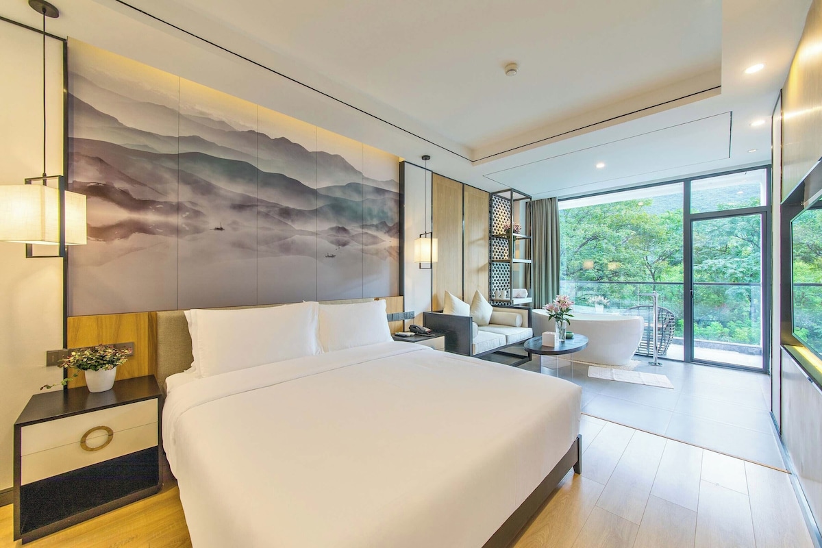 A modern hotel room with a large bed, a view of greenery through floor-to-ceiling windows, a wall-mounted artwork, and a small seating area with a table and chairs.