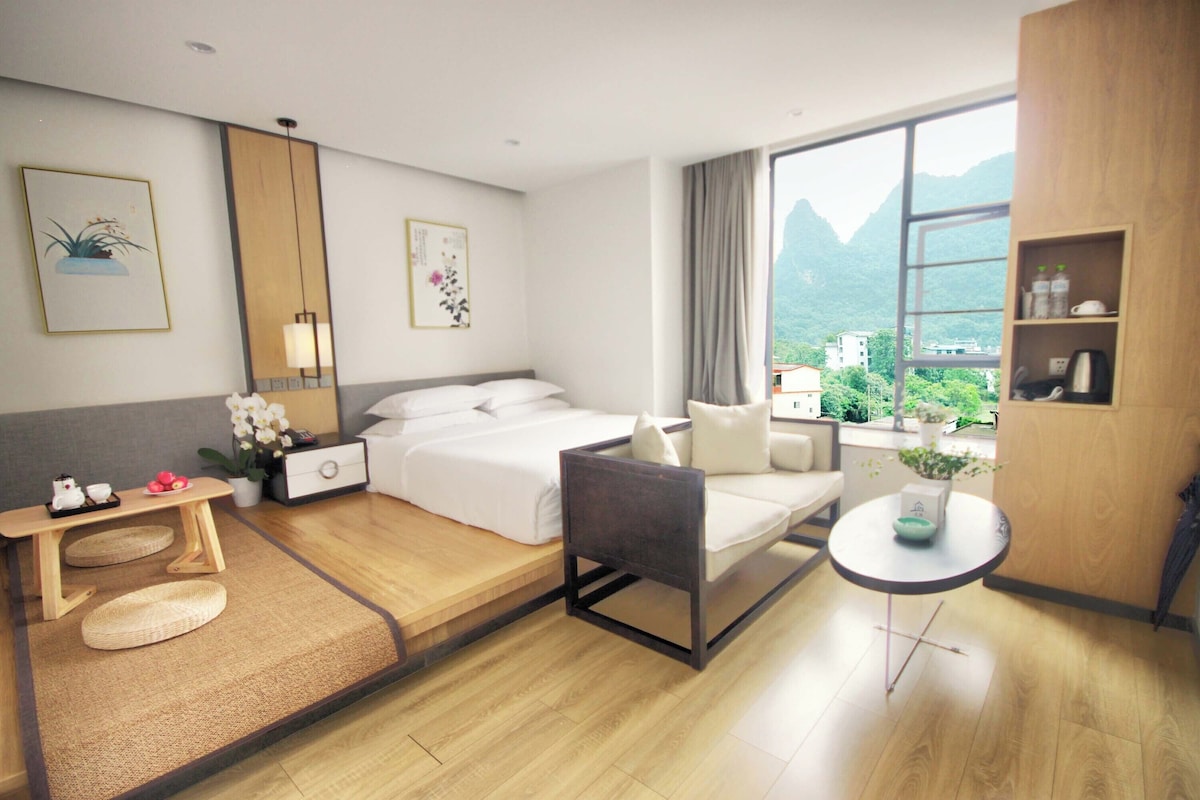 A hotel room with a bed, bedside table, chair, small table, and a view of mountains through a window.