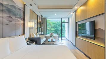 Habitación de hotel moderna con una cama grande, zona de estar con sofá y sillones, una mesita redonda, televisor de pantalla plana en la pared y un ventanal con vistas a zonas verdes.