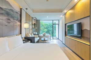 A modern hotel room with a large bed, a seating area with a sofa and armchairs, a small round table, a flat-screen TV mounted on the wall, and a large window offering a view of greenery outside. - Liangsu Shili Jiangshan Hotel (Guilin)