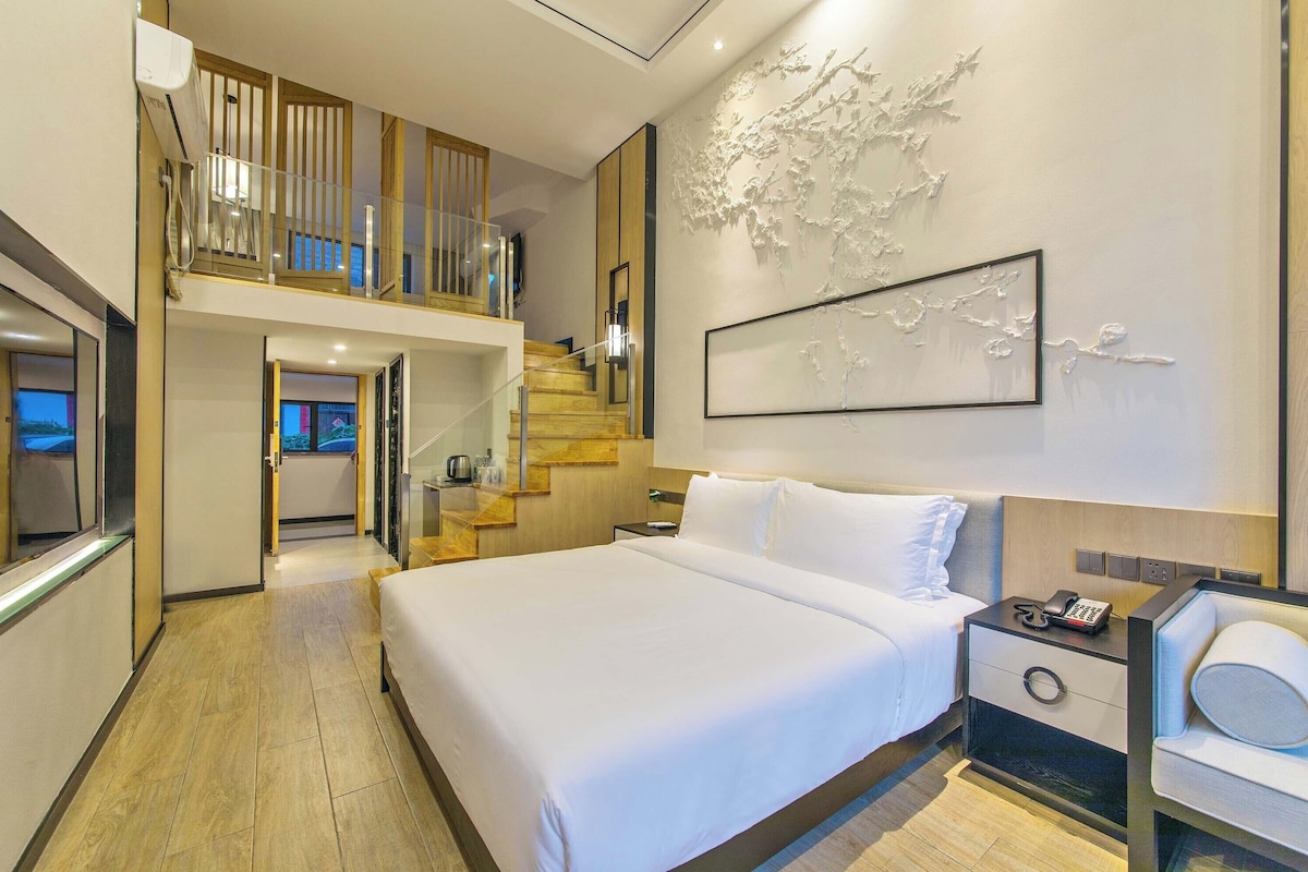 A hotel room with a large bed, a nightstand with a phone, a wall-mounted artwork, and a staircase leading to an upper level.