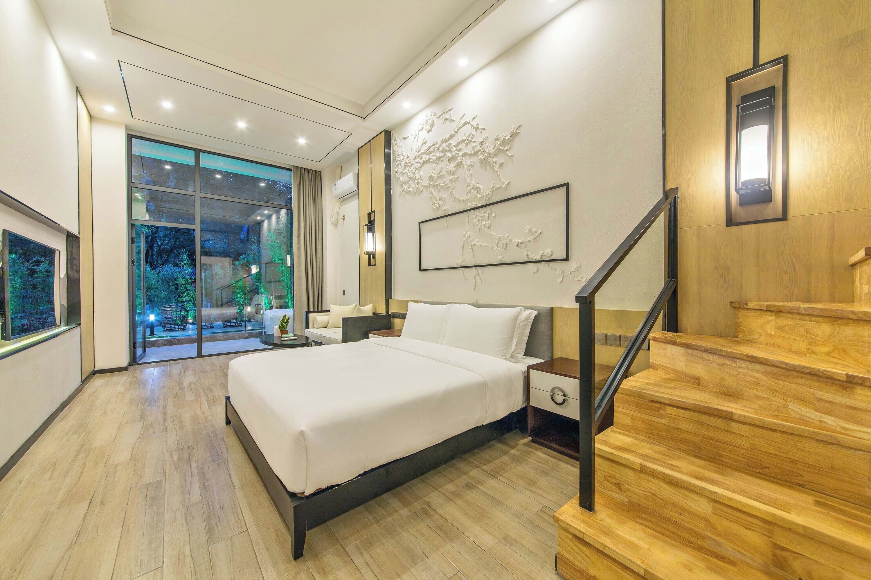 A modern hotel room with a large bed, a nightstand, a wooden staircase, and a view of a garden through a glass door.