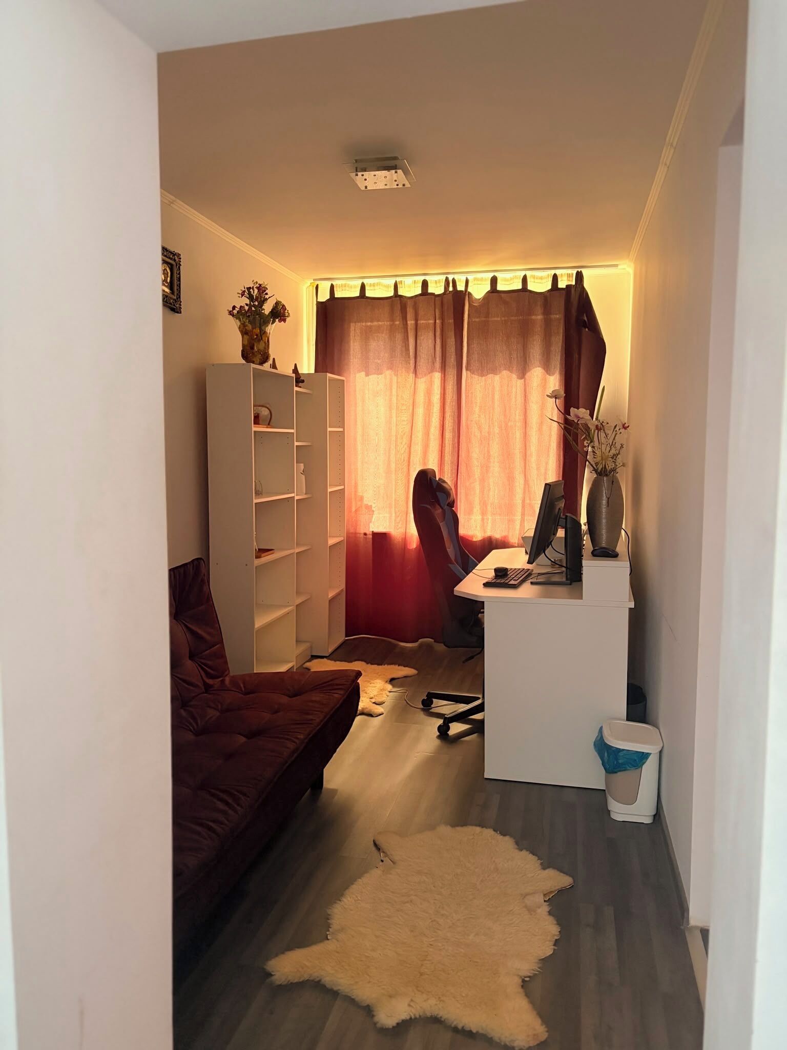 Un espace de vie compact comprenant un canapé, un bureau avec un ordinateur, une chaise, une bibliothèque et un vase décoratif.