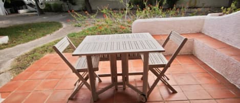 An outdoor patio with a wooden table and chairs, surrounded by a garden with various plants and a white wall.