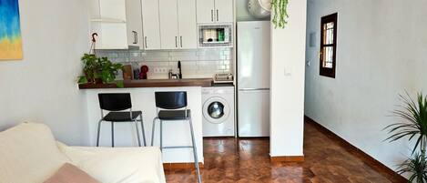Cocina compacta con armarios blancos, microondas, lavadora y una planta colgante.