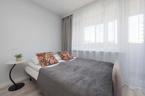 Ein modernes Schlafzimmer mit einem Bett, einem kleinen Nachttisch mit einer Pflanze und durchsichtigen Vorhängen am Fenster.