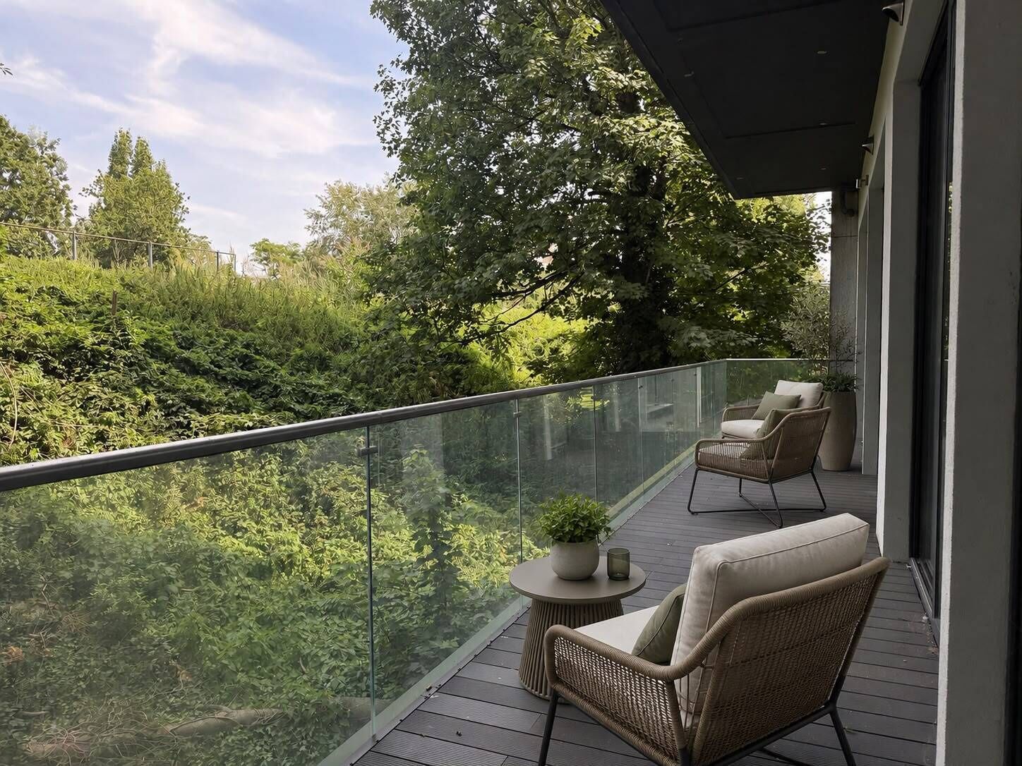 A balcony with wicker furniture, a small table with a plant, and a glass railing overlooking a lush green landscape.